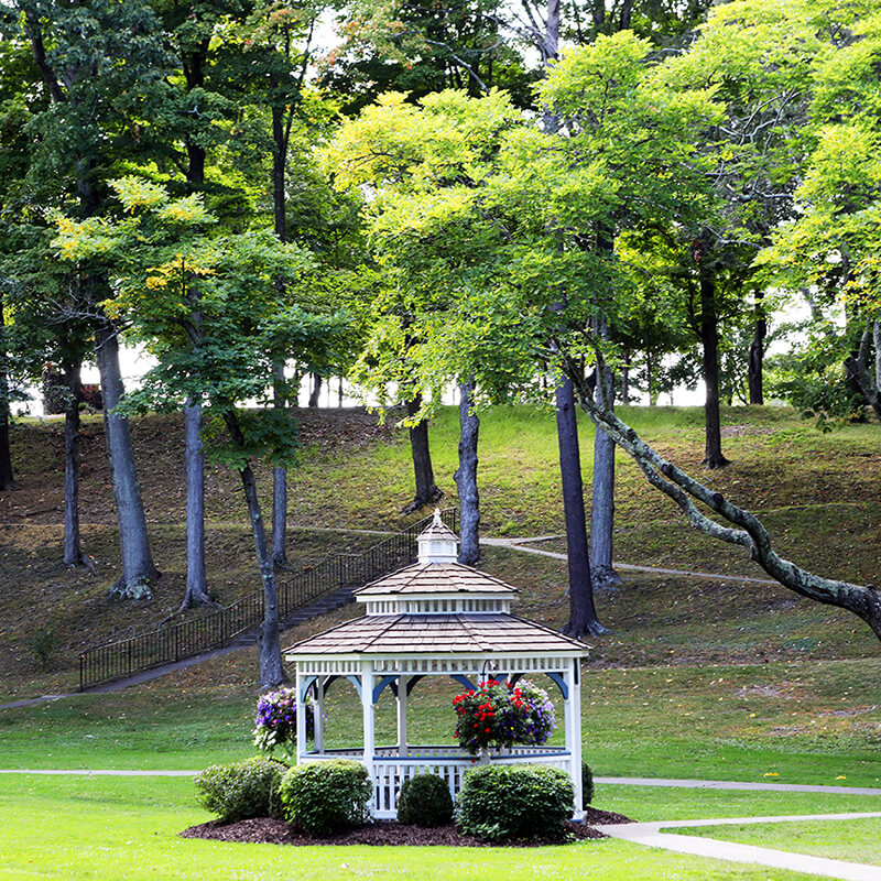 A gazebo in a public garden, surrounded by green grass and tall deciduous trees.